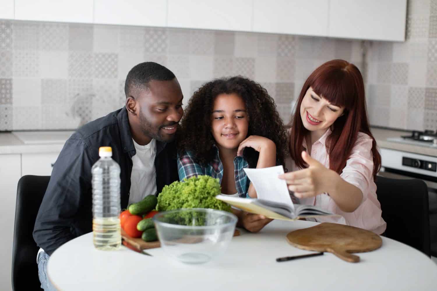 African man and Caucasian woman, teaching their teenage curly daughter cooking healthy food